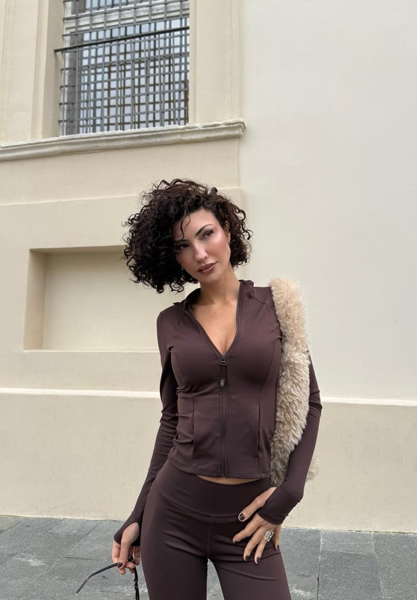 Kvinne med krøllete mørkt hår iført en tettsittende brun glidelåstopp og bukser, bærende en beige pelsjakke over den ene skulderen, stående på et fortau.