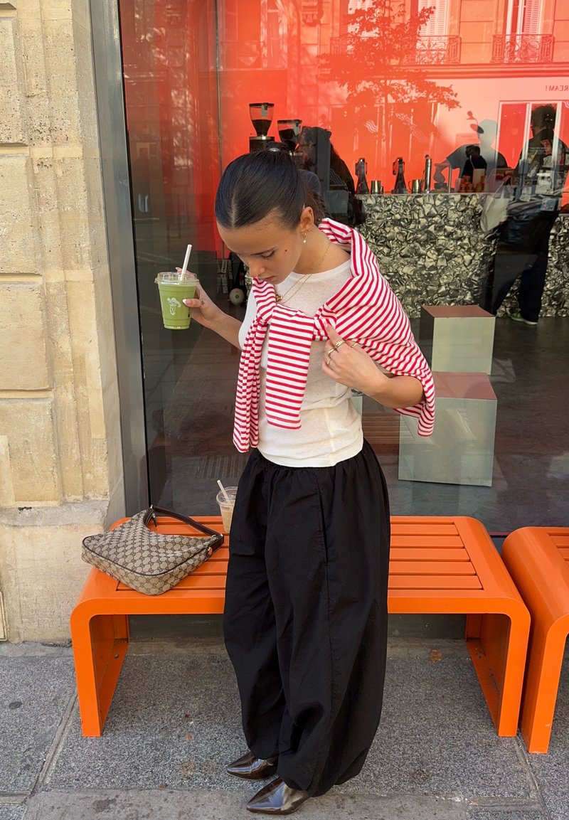 Striped red and white sweater tied around shoulders, white shirt, black pants, brown patterned bag, green drink, and orange bench.