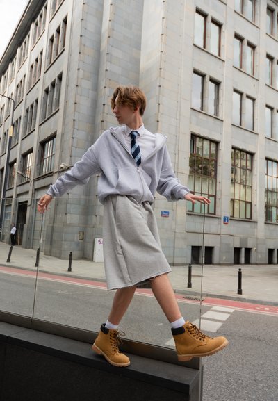 Sudadera gris con cremallera, camiseta blanca y corbata a rayas combinadas con una falda gris hasta la rodilla. Botas de encaje color marrón y fondo urbano con grandes ventanales.