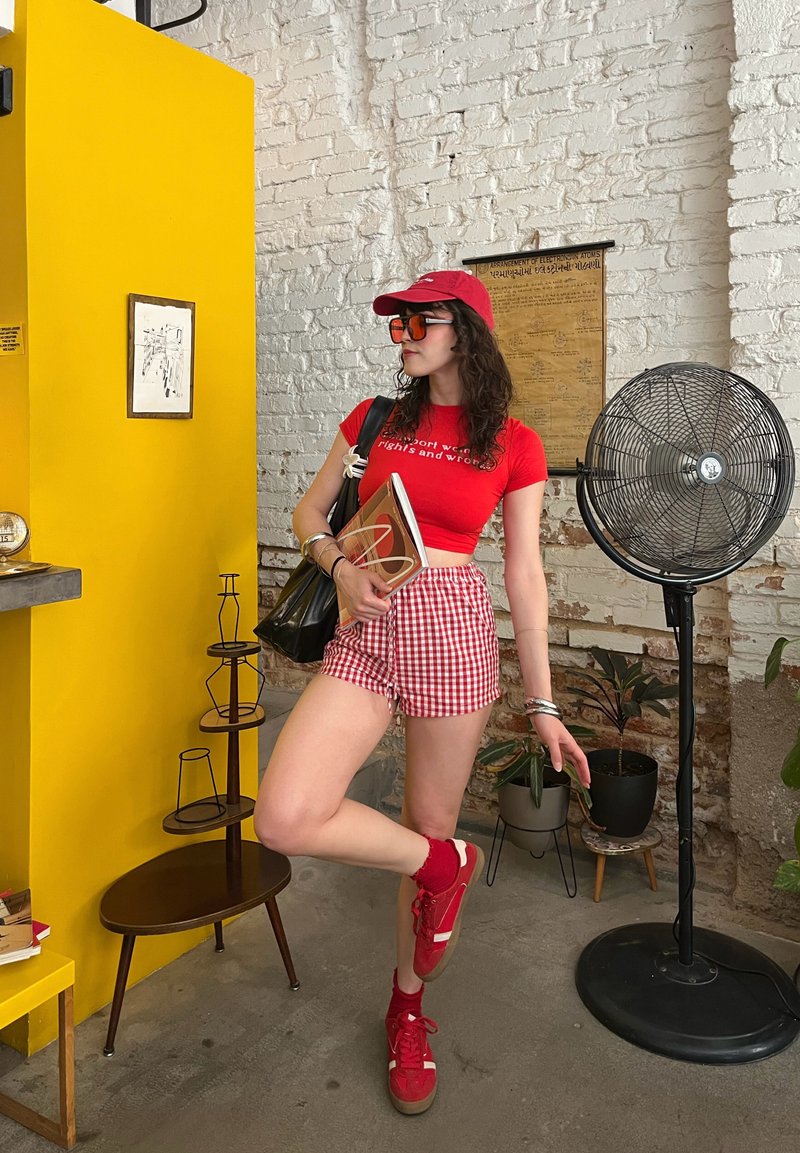 T-shirt rouge court avec texte, shorts à carreaux rouges, casquette rouge et lunettes de soleil. Baskets rouges associées à des chaussettes. Mur jaune en arrière-plan.
