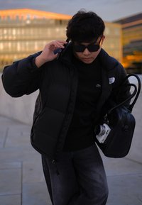 Μαύρο μπουφάν με ψηλό γιακά, συνδυασμένο με μαύρο πουλόβερ και σκούρα τζιν. Κρατάει μια μαύρη τσάντα και μια λευκή κάμερα. Φοράει σκούρα γυαλιά ηλίου.