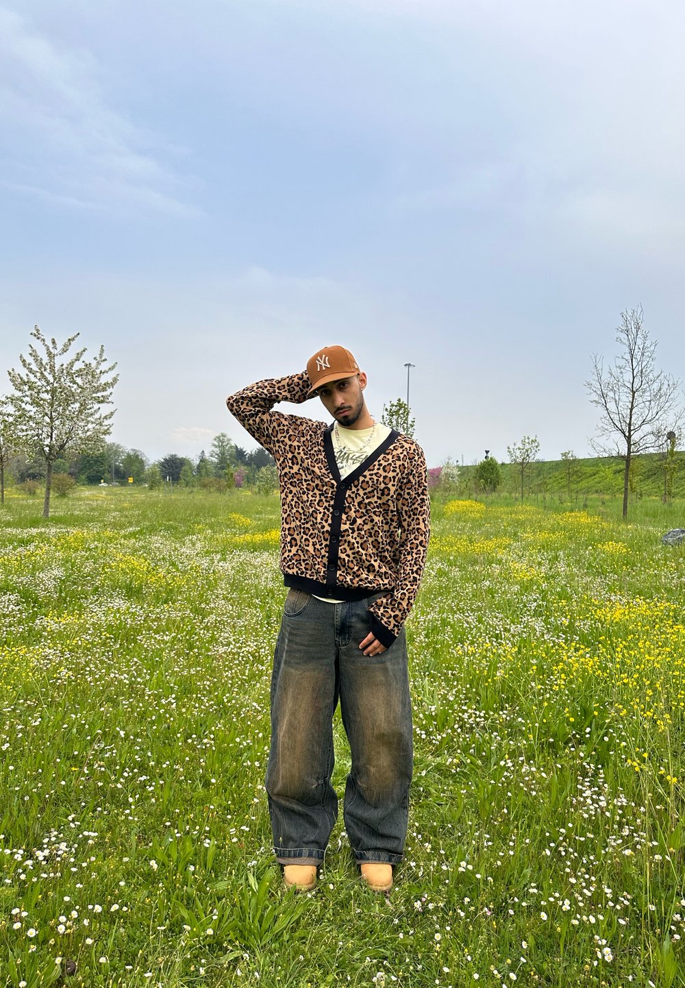 Κάρντιγκ από λεοπάρ σχέδιο με μαύρες λεπτομέρειες, χαλαρά γκρι τζην και καφέ καπέλο. Στέκεται σε ένα χλοερό πεδίο με αγριολούλουδα.