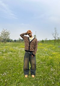 Κάρντιγκ από λεοπάρ σχέδιο με μαύρες λεπτομέρειες, χαλαρά γκρι τζην και καφέ καπέλο. Στέκεται σε ένα χλοερό πεδίο με αγριολούλουδα.