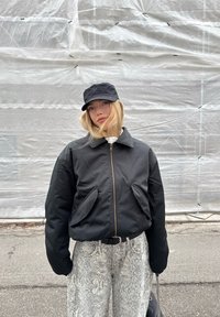 Sort bomberjakke med lynlås foran og to lommer, kombineret med grå vidde-ben-jeans med slangeprint og en sort kasket. Glat stoftekstur.