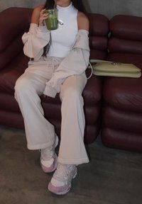 Personne assise sur un canapé en cuir marron en train de siroter une boisson verte, portant un haut blanc sans manches, un pantalon beige et des baskets, avec un sac vert à côté d'elle.