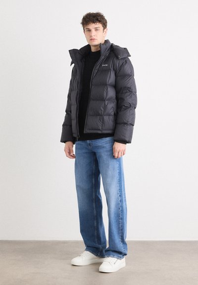 Chaqueta de plumas negra con capucha, con acolchado de rombos y cierre de cremallera. Lleva puesta sobre un suéter negro, con jeans rectos azules y zapatillas blancas.