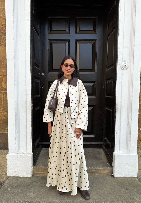 Femme portant une veste blanche à pois et une jupe avec un col et des chaussures marron, debout devant une porte noire à panneaux.