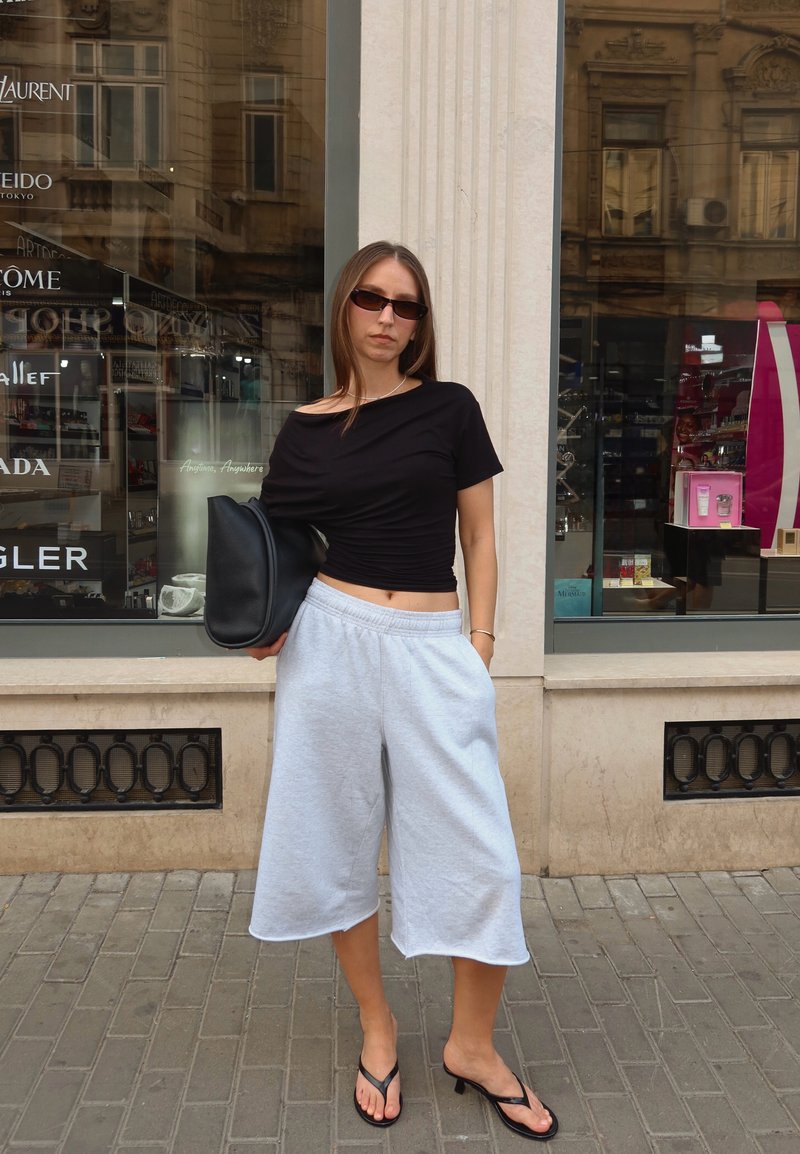 Mujer con gafas de sol, top negro, shorts anchos de color gris claro y sandalias negras de tacón, de pie en la acera de la ciudad sosteniendo un bolso negro bajo el brazo.