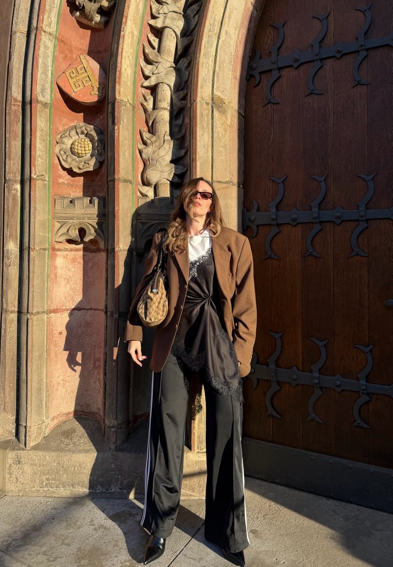 Femme portant un blazer marron, une robe en satin noire par-dessus une chemise blanche, un pantalon large noir et des lunettes de soleil, debout près d'un arc en pierre orné et d'une grande porte en bois.