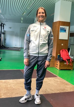 El atleta lleva una chaqueta con cremallera gris claro y mangas gris oscuro, combinada con pantalones de chándal a juego, y zapatillas blancas y negras. Se observa un ambiente de gimnasio.