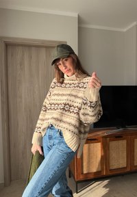 Femme en pull crème à motifs, jean bleu et casquette verte, posant à l'intérieur près d'un meuble TV en bois, tenant un vêtement vert.