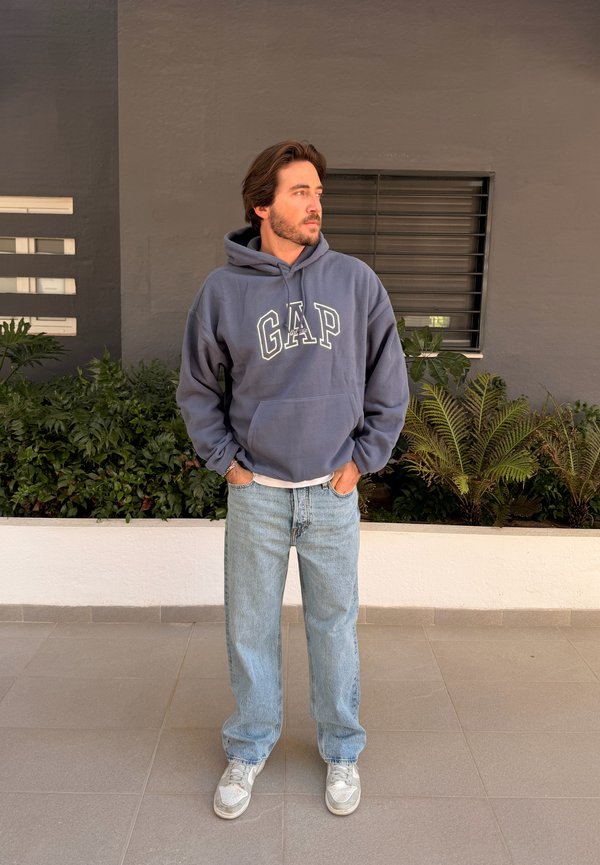 Νεαρός άνδρας με γενειάδα που φοράει μπλε φούτερ GAP, ανοιχτόχρωμα τζιν και λευκά αθλητικά παπούτσια, στέκεται με τα χέρια στις τσέπες κοντά σε φυτά και παράθυρο.