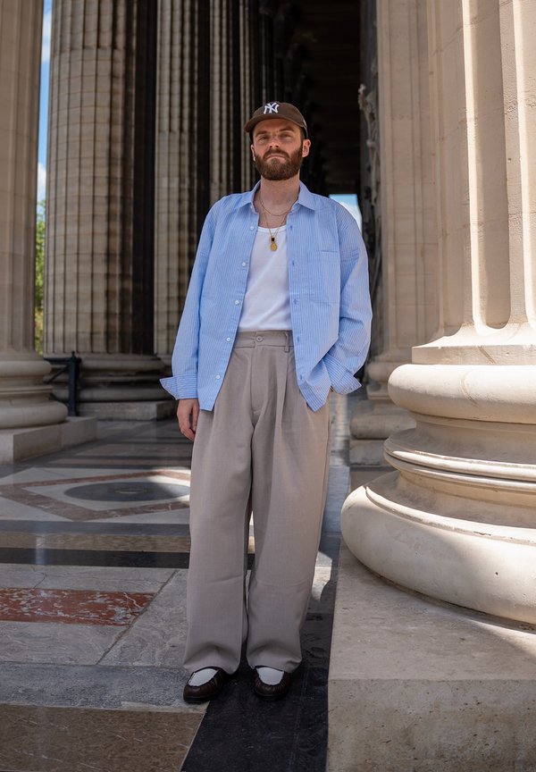 Homme barbu portant une casquette marron, une chemise à rayures bleues, un débardeur blanc, un pantalon large beige et des chaussures foncées se tient près de grandes colonnes en pierre.