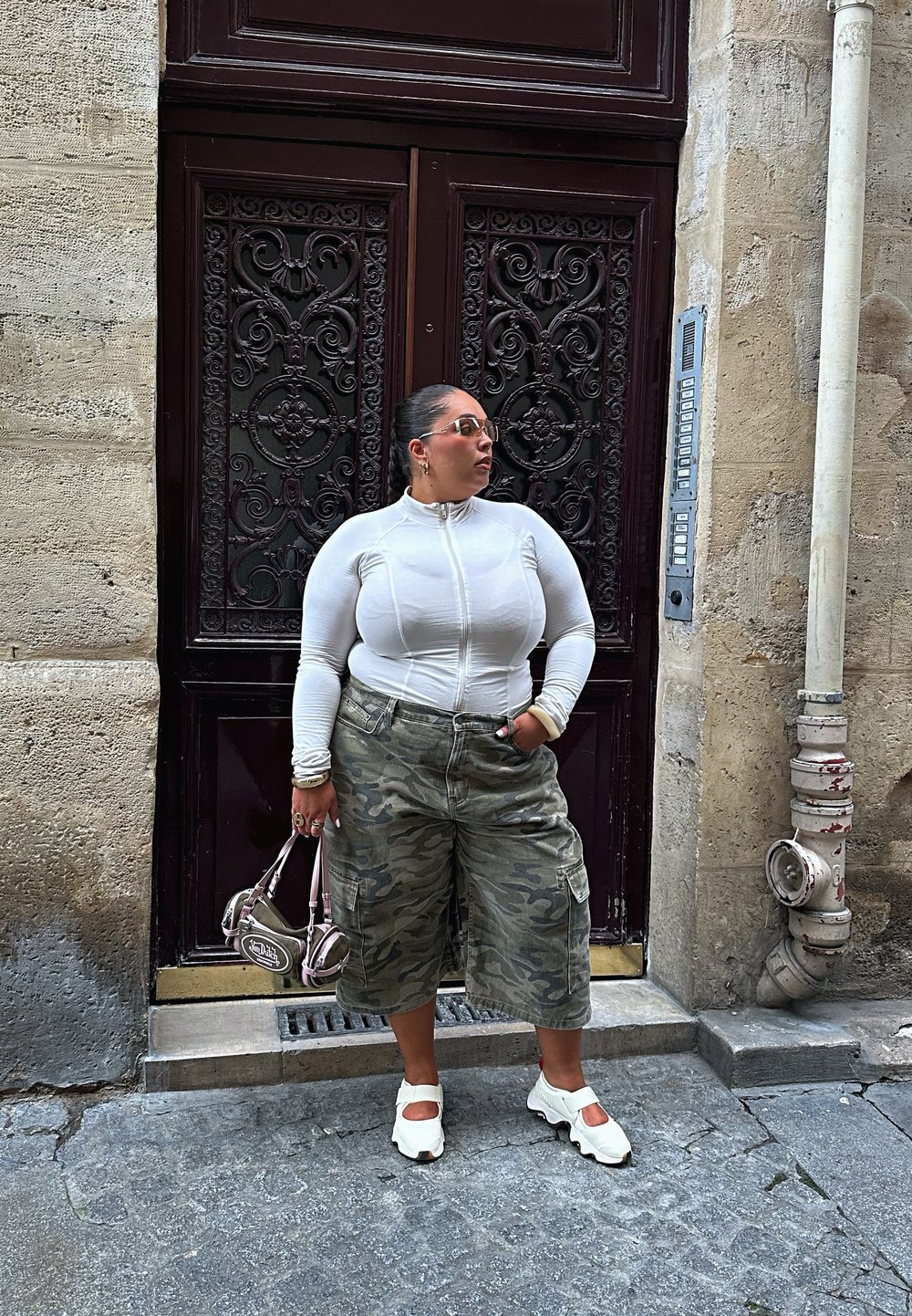 Kvinde i hvid zip-up trøje, camouflage brede shorts, hvide sko, der holder en lille håndtaske, stående ved en udsmykket mørk dør på en stenbelagt gade.