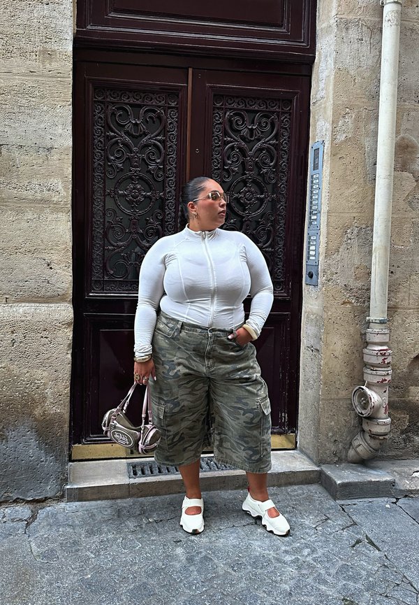 Mujer con parte superior blanca de cremallera, pantalones cortos de camuflaje de pierna ancha, zapatos blancos, sosteniendo un bolso pequeño, de pie junto a una puerta oscura ornamentada en una calle de piedra.