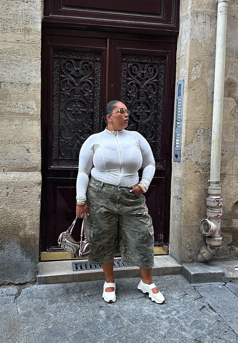 Donna in una maglietta bianca con zip, pantaloni corti larghi in camouflage, scarpe bianche, che tiene una piccola borsa a mano, in piedi accanto a una porta ornata scura su una strada di pietra.