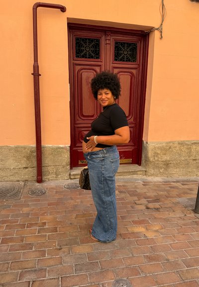 Top corto negro de manga corta, jeans anchos de tiro alto en azul claro y un bolso marrón. El fondo presenta una puerta marrón texturizada y un pavimento de ladrillo.