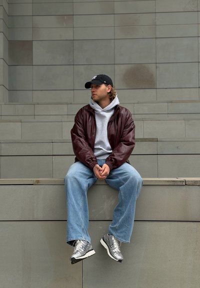 Joven vestido con ropa informal y gorra negra sentado en unos escalones de cemento con las manos entrelazadas, mirando hacia un lado frente a una pared con azulejos.