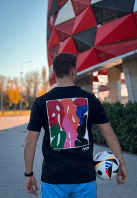 Μαύρο t-shirt με πολύχρωμο αφηρημένο σχέδιο στην πλάτη που περιλαμβάνει ροζ, πράσινα, κόκκινα, μπλε και μαύρα σχήματα. Ένα ποδοσφαιρικό μπαλάκι κρατιέται στο αριστερό χέρι.