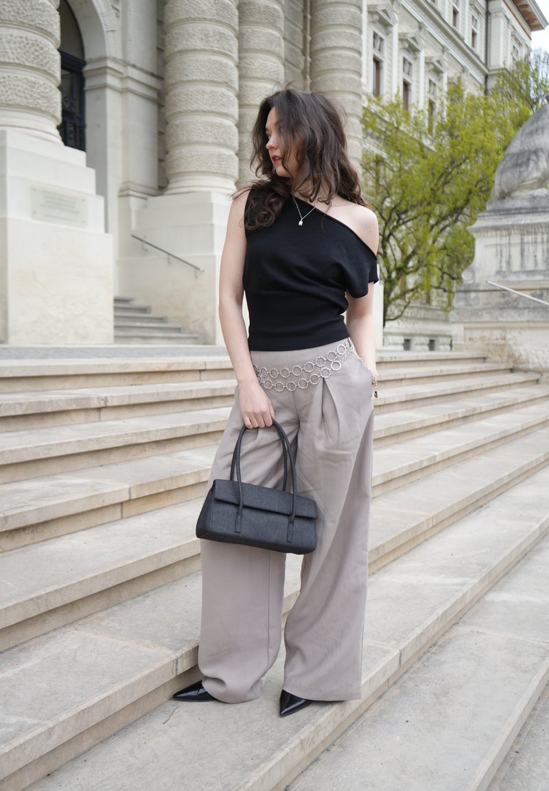 Γυναίκα με χαλαρό μαύρο τοπ και μπεζ φαρδιές παντελόνες, διακοσμημένη με ασημένια αλυσίδα μέσης και μαύρη τσάντα, στέκεται σε πέτρινα σκαλοπάτια σε εξωτερικό χώρο.