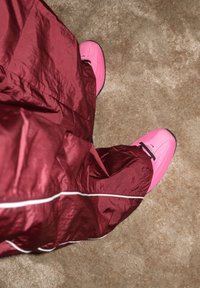 Chaussures de sport roses avec des lacets noirs, partiellement couvertes par un pantalon bordeaux brillant et ample avec une bande blanche, posées sur un tapis beige.