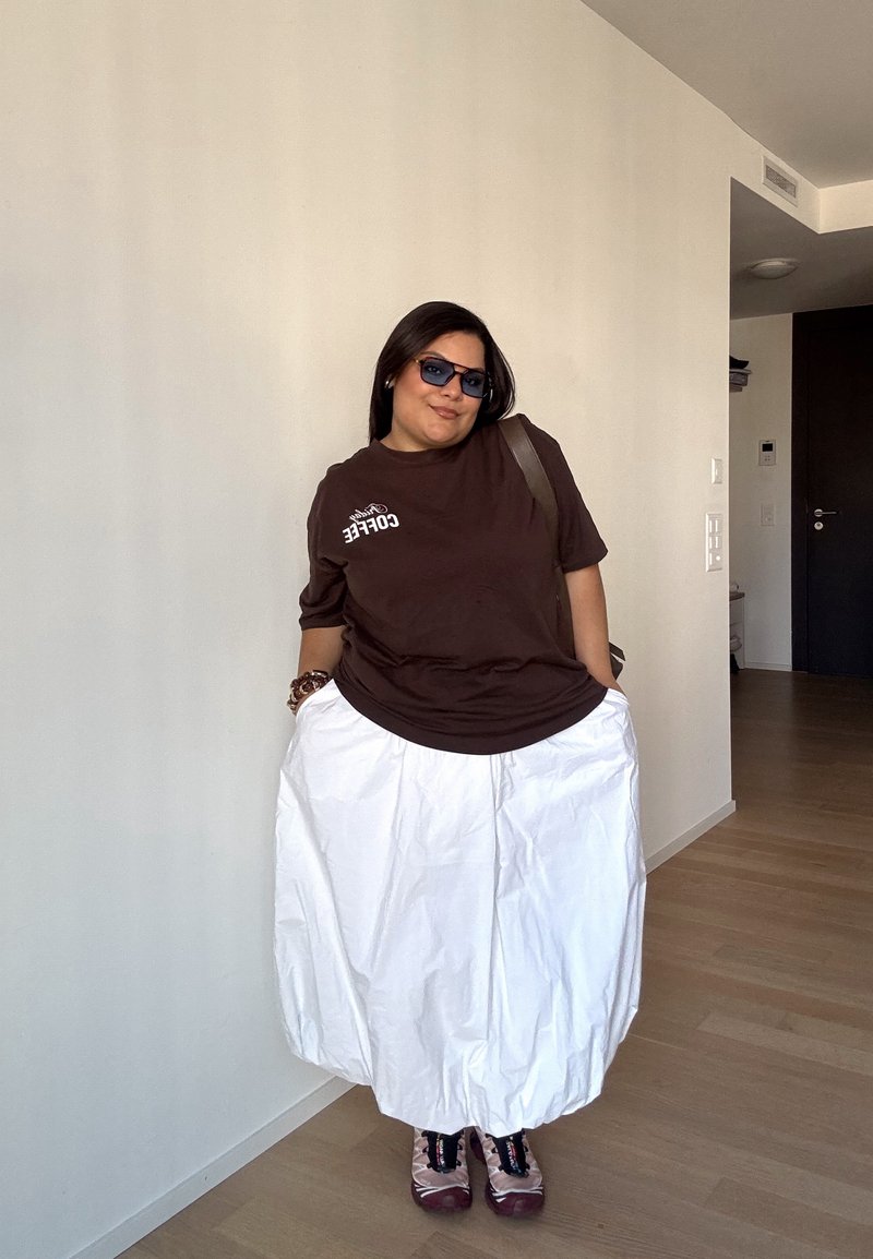 Mulher a usar óculos de sol, uma camisa castanha com temática de café, uma saia larga branca e sapatilhas, em pé no interior com as mãos nos bolsos.