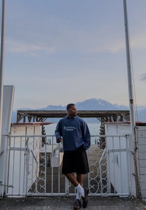 Hombre con sudadera azul marino y pantalones cortos negros apoyado en una puerta blanca junto a un muelle en la costa con montañas y agua tranquila de fondo al anochecer.