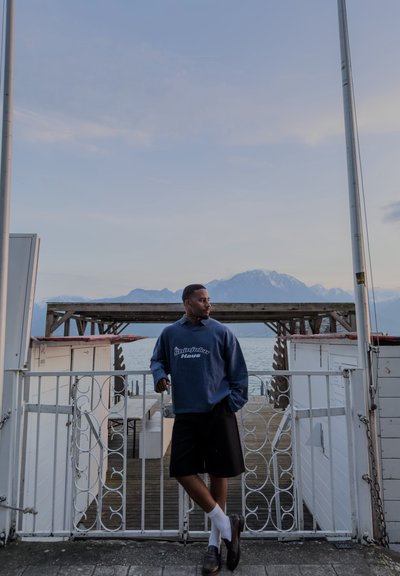 Hombre con sudadera azul marino y pantalones cortos negros apoyado en una puerta blanca junto a un muelle en la costa con montañas y agua tranquila de fondo al anochecer.