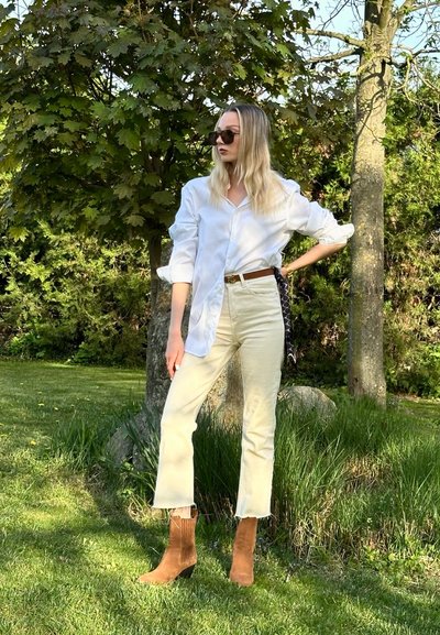Camisa blanca de botones, jeans de talle alto en color beige claro, botas tobilleras de punto marrón y un cinturón con estampado. Ambiente exterior con vegetación.