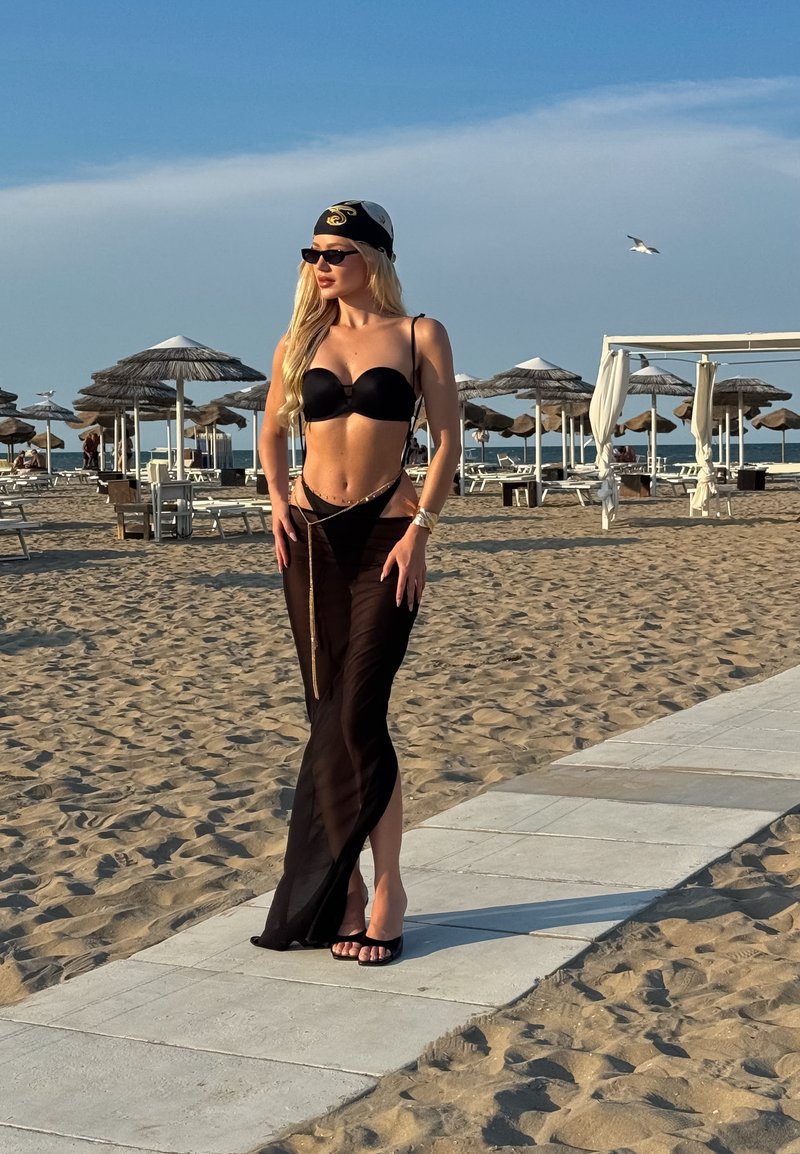 Femme en bikini noir et jupe légère transparente debout sur un chemin de plage avec des parasols et des chaises longues en arrière-plan.
