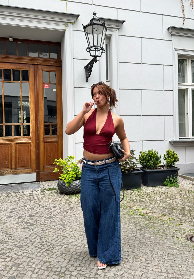 Jeune femme en top rouge à bretelles et jean large debout dans une rue pavée, tenant un sac noir et léchant une sucette près d'une porte en bois et de plantes en pot.