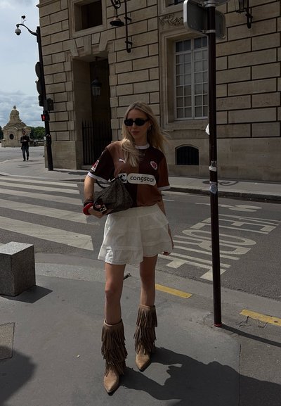 Jersey deportivo marrón con acentos blancos, falda blanca con volantes en capas, botas de ante beige con flecos y bolso estampado, llevado con gafas de sol oscuras.