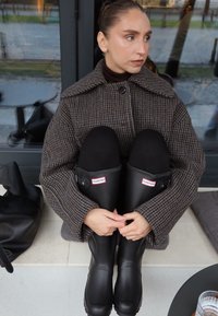 Femme en manteau à carreaux et leggings noirs, assise les genoux relevés, portant des bottes de pluie Hunter noires devant une porte en verre.