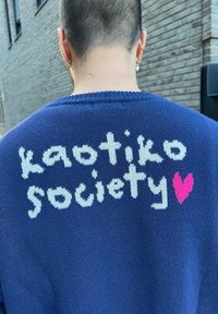 Pullover navy con testo lavorato "kaotiko society" in bianco e un cuore rosa. La texture è morbida, con colletto a coste e una vestibilità comoda.