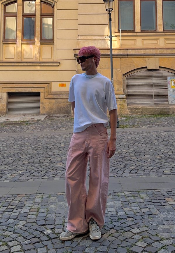 Persona con capelli rosa e occhiali da sole, che indossa una maglietta bianca, pantaloni rosa a gamba larga e scarpe da ginnastica bianche, in piedi su una strada lastricata.