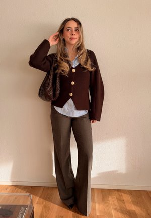 Mujer de pelo largo con cárdigan marrón con botones dorados, camisa azul clara, pantalones marrones, sosteniendo un bolso tejido marrón, de pie en interiores.