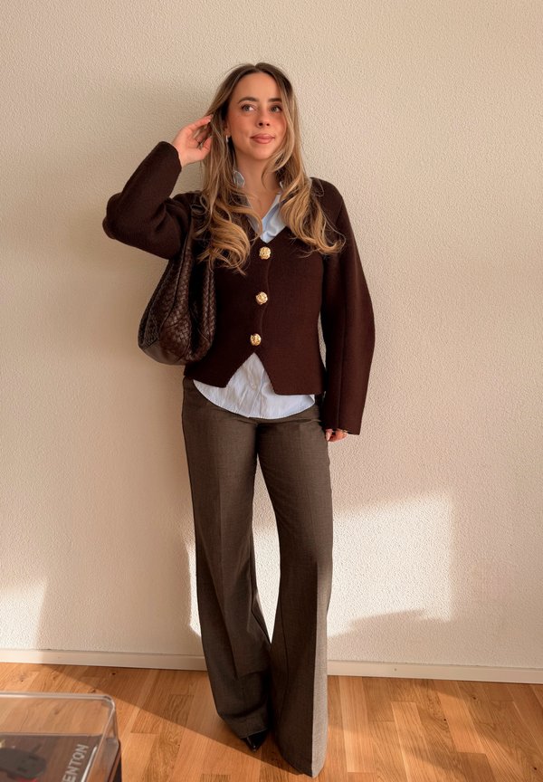 Žena s dlhými vlasmi, oblečená v hnedom kardigane so zlatými gombíkmi, svetlomodrej košeli, hnedých nohaviciach, drží hnedú tkanú kabelku, stojí v interiéri.