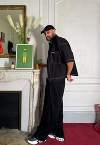 Μαύρο υπερμέγεθες πουκάμισο κοντό μανίκι με λευκές ραφές, μαύρο φαρδύ παντελόνι και φαρδιά αθλητικά παπούτσια με σχέδια. Λευκό μάρμαρο τζάκι και πολύχρωμα έργα τέχνης περιλαμβάνονται.