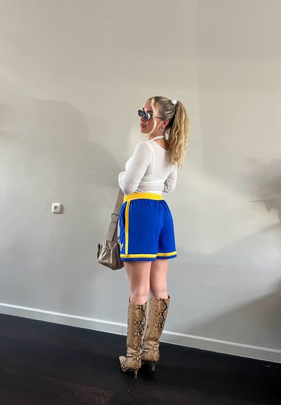 Pantalones cortos deportivos azules con acentos amarillos, combinados con una camisa blanca de manga larga y botas altas de serpiente hasta la rodilla. Bolsa de mano marrón, gafas de sol puestas.
