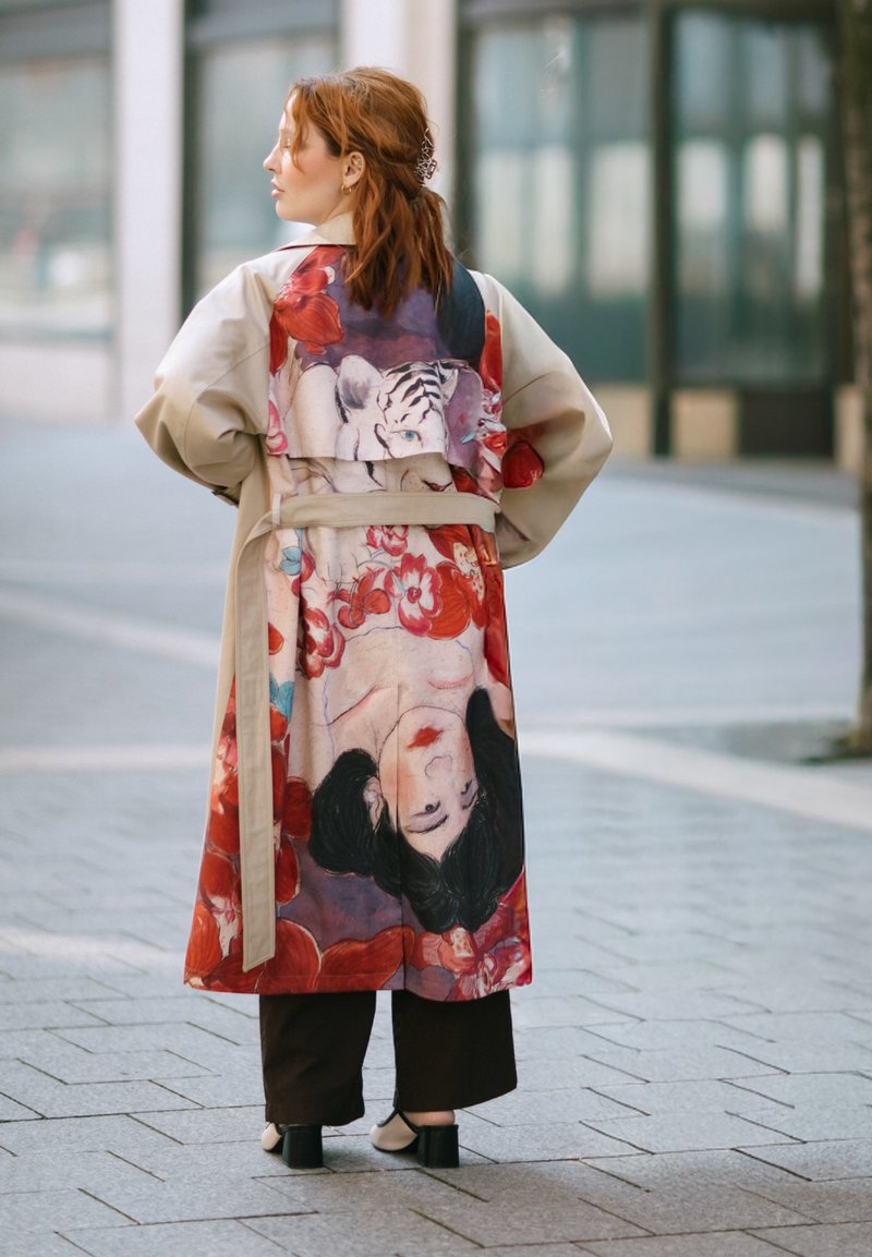 Femme aux cheveux roux se tient dans une rue de la ville portant un manteau beige avec un imprimé artistique coloré d’un visage de femme, d’un tigre et de fleurs rouges.