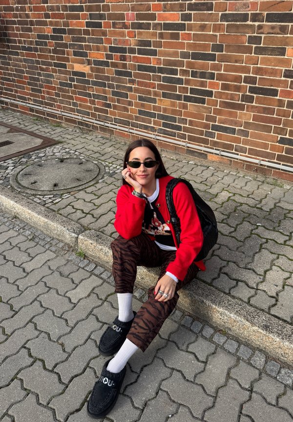 Cardigan roșu peste un tricou negru cu imprimeu grafic, pantaloni maro cu model zebra, șosete albe și pantofi negri. Accesoriile includ inele și ochelari de soare.
