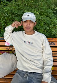 Weißes geripptes Langarmshirt mit "ELLESSE"-Logo, graue Jeans und eine weiße Nike-Cap. Schlüsselbund mit sichtbaren Schlüsseln an der Hüfte. Grüner Hintergrund.