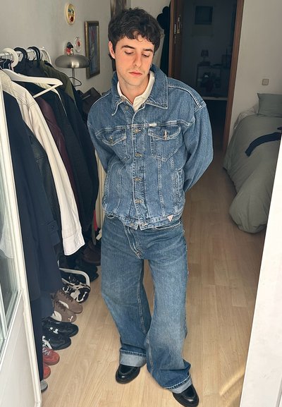 Chaqueta de denim y jeans de pierna ancha en azul medio, con cierre de botones y bolsillos en el pecho. Zapatos negros con acabado brillante.