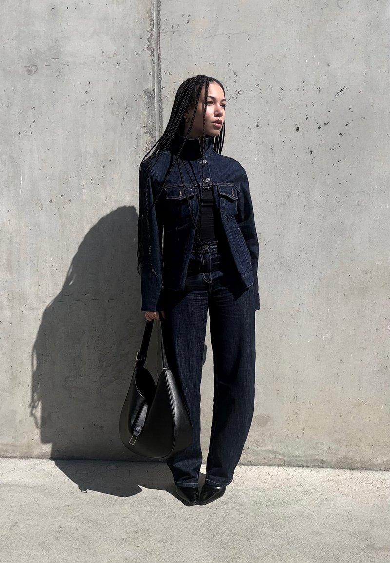 Woman with long braids wearing a black denim jacket and pants, holding a black handbag, standing against a concrete wall in sunlight.