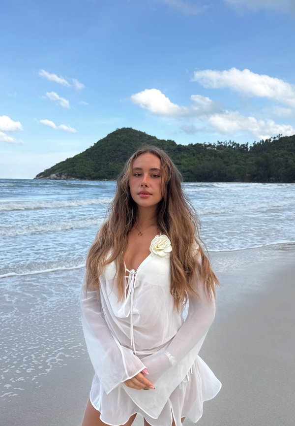 Vit genomskinlig överdel med långa ärmar och dragsko i halsringningen, prydd med en vit ros på vänster sida, strand i bakgrunden.