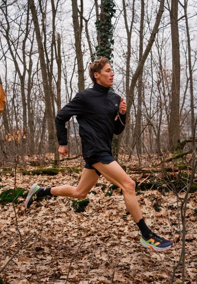 Chaqueta ligera negra, pantalones cortos de correr y zapatillas New Balance coloridas. Corredor en un entorno forestal con hojas secas bajo los pies.