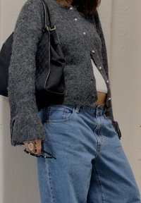 Henkilö, joka käyttää harmaata napitettavaa neuletakkia ja vaaleansinisiä farkkuja, pitää käsissään mustia aurinkolaseja ja kantaa mustaa käsilaukkua olkapäällä.