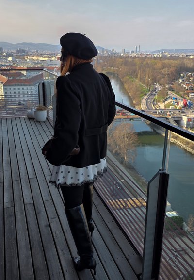 Mujer con abrigo negro, boina y botas, de pie en un balcón con vistas a un río, paisaje urbano y carreteras durante el día.