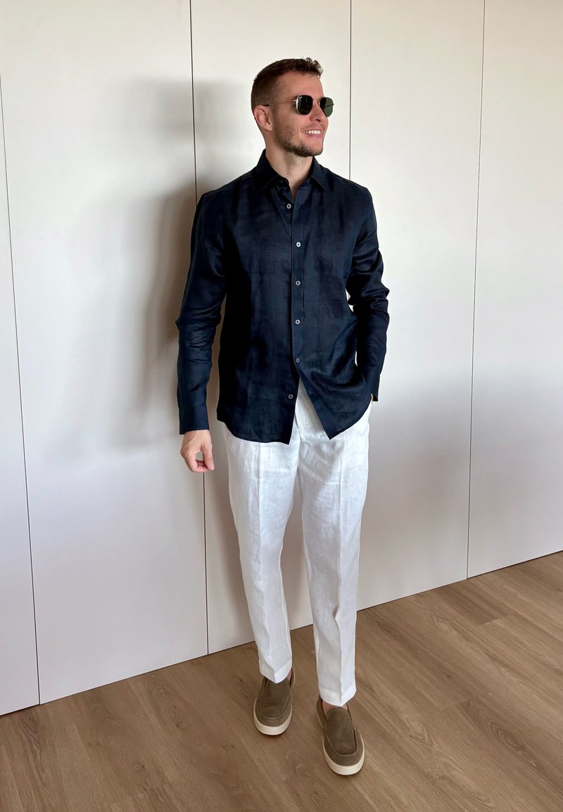 Homme portant une chemise bleu foncé à boutons, un pantalon blanc, des chaussures marron sans lacets et des lunettes de soleil, debout sur un sol en bois contre un mur blanc.