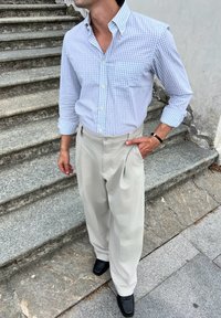 Camicia a quadri blu chiaro e bianca, infilata in pantaloni plissettati crema, abbinata a scarpe in pelle nera, in piedi su gradini di pietra.
